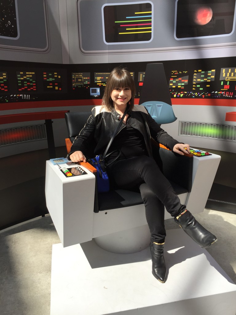 Image of Elissa Smith sitting in the captain's chair from Star Trek. She is wearing a leather jacket, jeans and high heels.